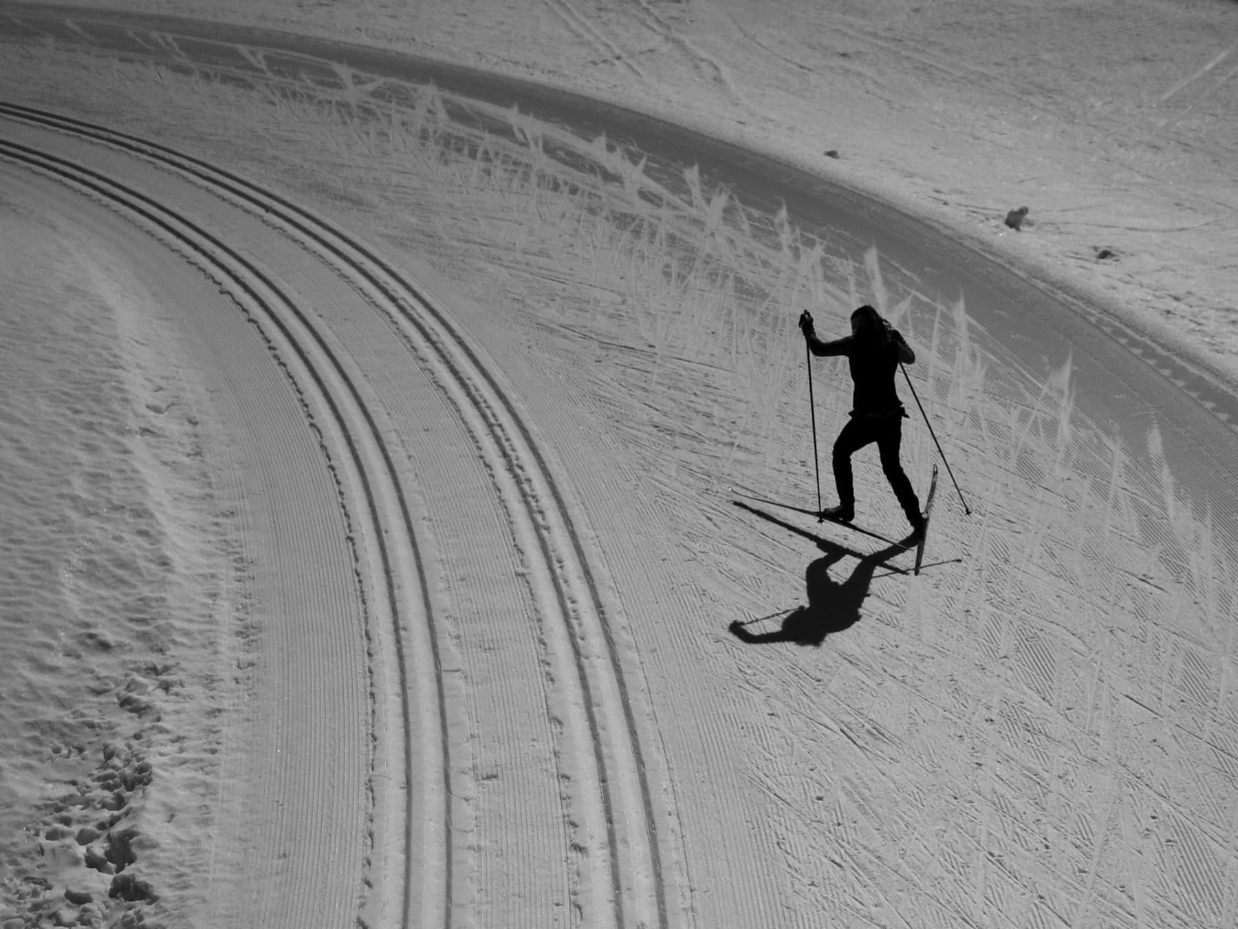 Black-and-white aerial view of a lone skier climbing beside curving classic tracks